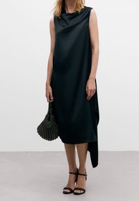 Robe noire sans manches avec un col drapé et un ourlet asymétrique. Accessoirisée avec un sac à main en perles et des sandales noires à lanières.