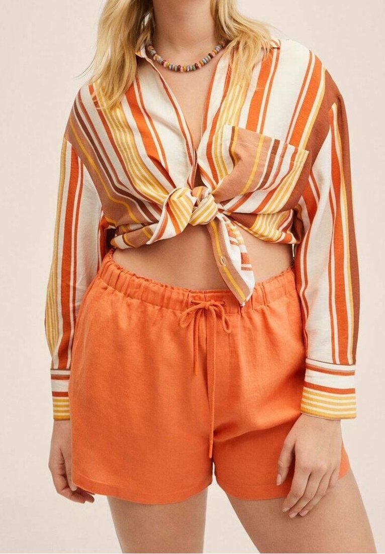 Femme porte un short orange à cordon et une chemise à manches longues boutonnée avec des rayures verticales orange, jaune et blanches nouée à la taille.