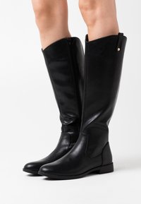 Black leather knee-high boots with a smooth texture, side zipper, and flat sole. Features a small metallic accent near the top.