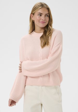 Mladá žena s blond vlasmi v ľahkom ružovom pletenom svetri a krémových nohaviciach stojaca pred jednoduchým bielym pozadím.