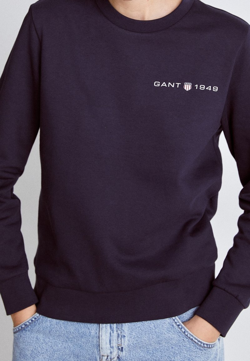 Sweat-shirt bleu marine foncé à manches longues avec logo "GANT 1949" et emblème bouclier sur le côté gauche de la poitrine, porté avec un jean bleu clair, mains dans les poches.