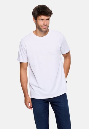 Hombre con camiseta blanca de manga corta y diseño sutil, y vaqueros azules, de pie con una mano en el bolsillo.