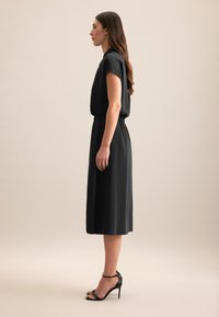 Schwarzes Midi-Kleid mit kurzen Ärmeln und taillierter Taille. Glatter Stoff, lockere Passform und kombiniert mit schwarzen High-Heels.