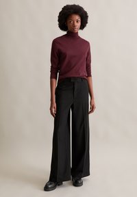 Bordeaux coltrui, aangesloten model, gecombineerd met wijde zwarte broek, gladde stof en massieve zwarte schoenen met een gestructureerde zool.