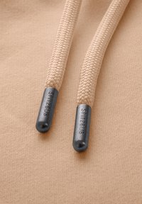 Beige gevlochten trekkoorden met metalen uiteinden gemarkeerd "SPITZBUB" op een zachte, gestructureerde achtergrond met een gladde afwerking.