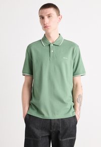 Žalias polo marškinėlis, pagamintas iš medvilnės, su trimis mygtukais priekyje, kontrastinga balta apdaila ant apykaklės ir rankovių, bei maža logotipas.