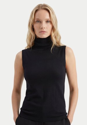 Femme aux cheveux blonds portant un haut noir sans manches à col roulé, debout devant un fond clair uni, mains dans les poches.