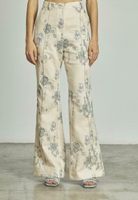 Vrouw die hoge, crèmekleurige, wijde broek draagt met blauwe bloemenbroderie en lichtblauwe sandaaltjes met bandjes, staande op een gladde vloer.