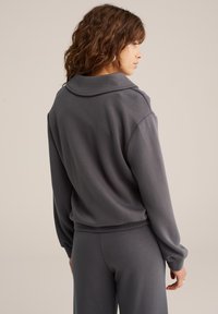 Mujer con pelo rizado vistiendo un sudadera holgada de color gris oscuro y pantalones a juego, de pie con la espalda vuelta, fondo neutro.