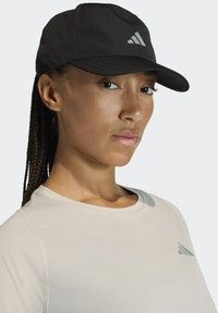 Gorra de béisbol negra con visera curva y un logo de tres franjas blancas. Se lleva con una camiseta deportiva gris claro con cuello redondo.