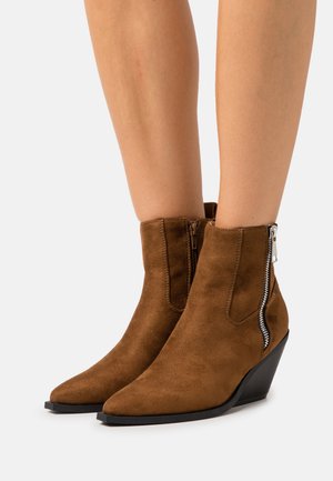 Botas de ante marrón hasta el tobillo con punta afilada y tacón de cuña. Cuentan con una cremallera lateral con un tirador metálico, que añade textura y detalle.