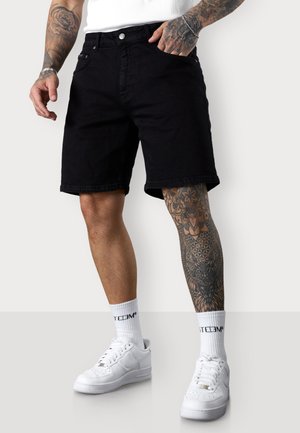 Schwarze Denim-Shorts mit geradem Schnitt, Knopfverschluss und vorderen Taschen. Getragen mit weißen Turnschuhen und sichtbaren gemusterten Tattoos am Bein.