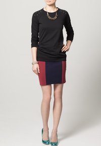 Top nero a maniche lunghe con spalline borchiate, abbinato a una gonna color block in blu scuro e bordeaux. Indossato con tacchi alti color teal.