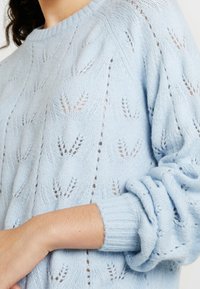 Maglione azzurro chiaro lavorato a maglia con un motivo in pizzo, dotato di polsini a costine e scollatura rotonda. La texture è morbida e leggera.