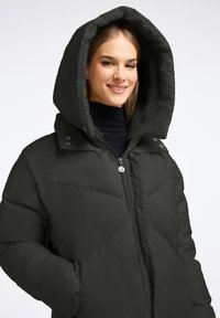 Donkergroene pufferjack met grote capuchon, ritssluiting en geborduurd patroon. Fleece voering zichtbaar rond de capuchon. Zakken aan de zijkanten inbegrepen.