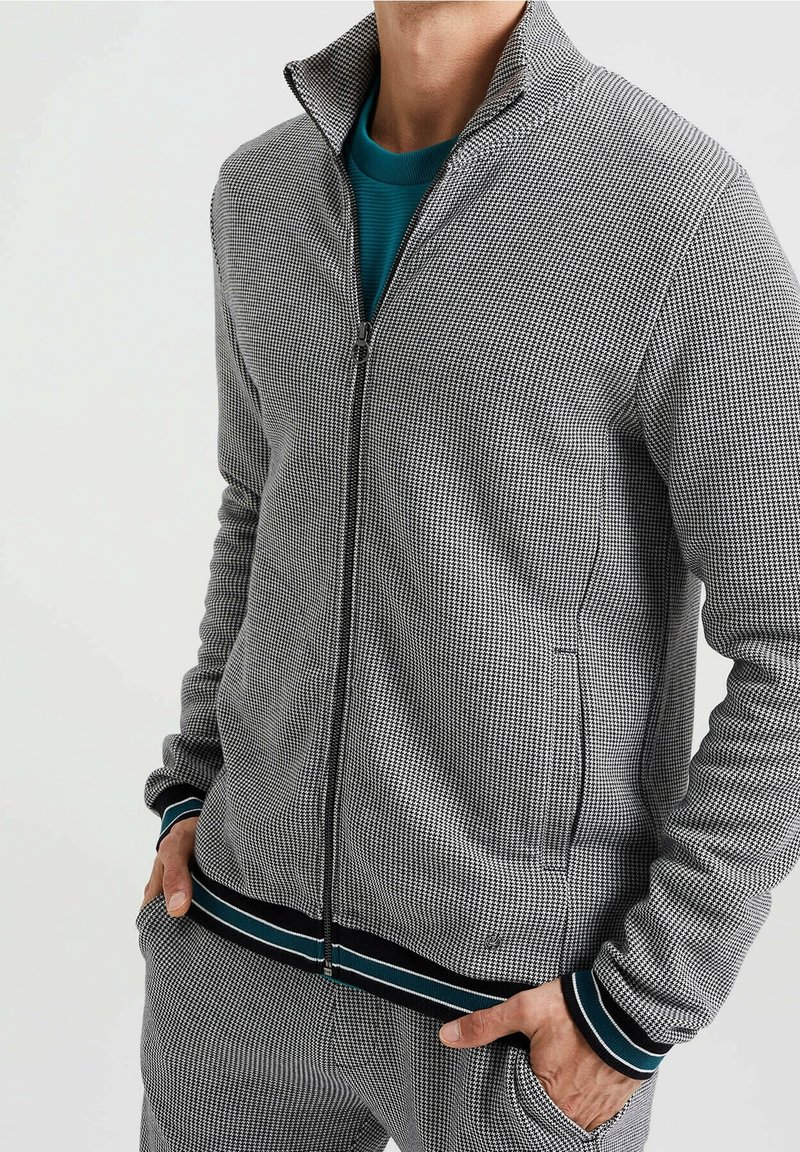 Grå och svart rutig zip-jacka med hög krage, med randiga detaljer på ärmsluten och fållen, tillverkad av strukturerad tyg.