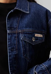 Chaqueta de denim azul con un collar clásico, costuras visibles, un bolsillo delantero con botón a presión y una etiqueta con el logotipo de Calvin Klein.