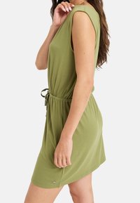 Lyhythihainen, V-pääntie mekko oliivinvihreänä, valmistettu pehmeästä kankaasta. Varustettu joustavalla vyötäröllä, jossa on nyöri ja hillitty logo helman lähellä.