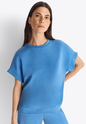 Sweatshirt bleu surdimensionné avec des manches courtes retroussées, texture lisse et col rond. Associé à un bas assorti.