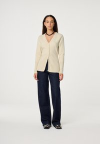 Beige cardigan with a V-neck and button closure, paired with dark blue wide-leg jeans. Smooth texture and fitted design enhance silhouette.