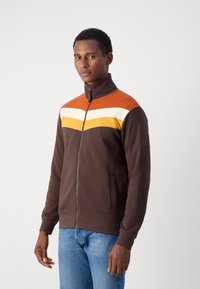 Braune Zip-Jacke mit orangefarbenen, weißen und gelben Streifen über der Brust. Hat einen hohen Kragen und Seitentaschen. Weicher Stoff.
