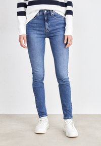 Skinny jeans in mediumblauwe denim, met een hoge taille, klassieke vijfzakkenontwerp en subtiele vervaging. Gepaarde met witte sneakers.