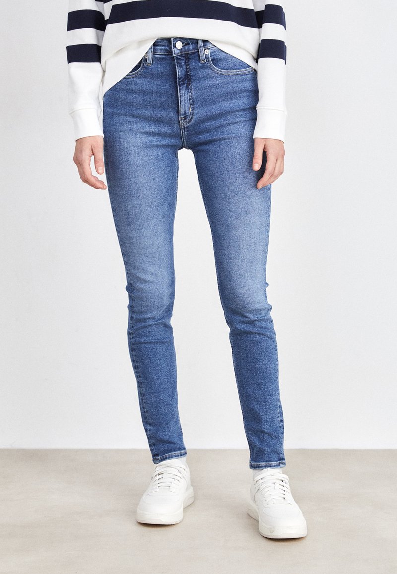 Skinny jeans in mediumblauwe denim, met een hoge taille, klassieke vijfzakkenontwerp en subtiele vervaging. Gepaarde met witte sneakers.