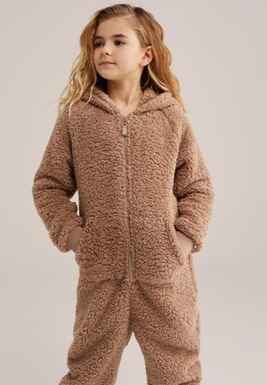 Junges Mädchen, das einen braunen Fleece-Overall mit Reißverschluss und Kapuze trägt und mit den Händen in den Taschen vor einem schlichten Hintergrund steht.