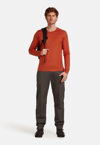 Mies, joka on pukeutunut oranssipunaiseen pitkähiharaiseen paitaan ja harmaisiin housuihin, seisoo pidellen mustan repun kantohihnaa toisen olkapään yli valkoista taustaa vasten.