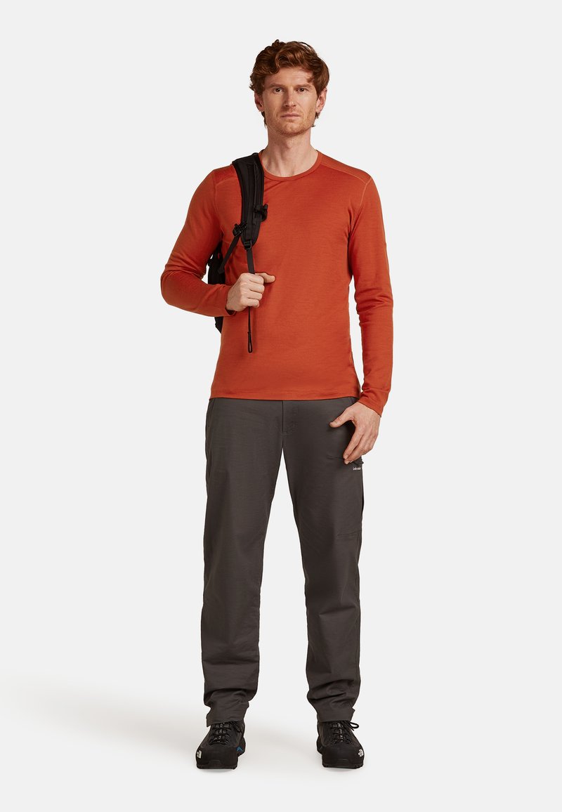 Mies, joka on pukeutunut oranssipunaiseen pitkähiharaiseen paitaan ja harmaisiin housuihin, seisoo pidellen mustan repun kantohihnaa toisen olkapään yli valkoista taustaa vasten.