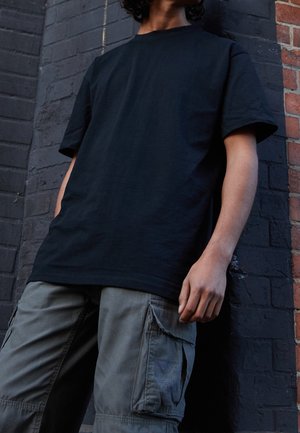 Sort bomuld T-shirt med korte ærmer kombineret med grå cargobukser med flere lommer og et struktureret stof mod en murstensvæg.