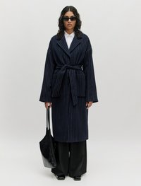 Navy gestreepte jas met wijde mouwen, een strikriem en twee voorzakken. Gecombineerd met zwarte wijde broek en een elegante zwarte tote bag.