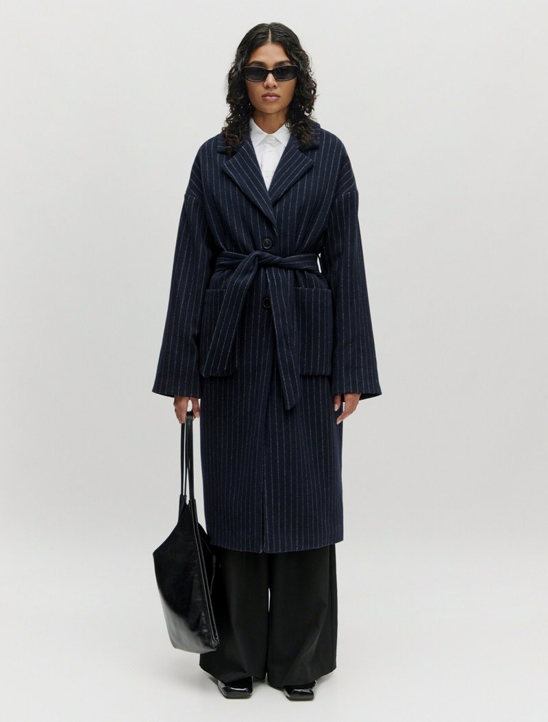 Navy gestreepte jas met wijde mouwen, een strikriem en twee voorzakken. Gecombineerd met zwarte wijde broek en een elegante zwarte tote bag.