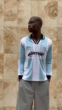 Maglia a maniche lunghe in blu chiaro e bianco con dettagli neri; presenta un colletto e un logo sul davanti; indossata con pantaloni larghi grigi chiari.