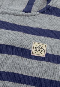 Grå strikket sweater med marineblå vandrette striber. Har et vævet mærke i tan med sort skrift og krydsede elementer.