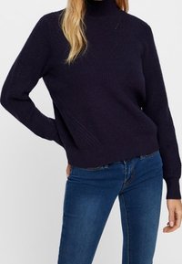 Merensininen neulottu neulepaita korkealla kauluksella, jossa on tekstuuri- ja resoriset hihansuut. Yhdistetty tummansinisiin farkkuihin. Minimalistinen tyyli.