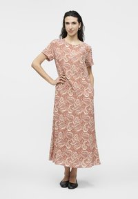 Vrouw draagt een jurk met korte mouwen en een enkel-lengte met een rood-wit bloemenpatroon, staand met één hand op haar heup en zwarte platte schoenen.