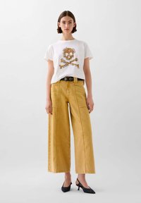 Wit T-shirt met een gele en zwarte schedelafbeelding, gecombineerd met hoog uitgesneden, wijde gele broek en marineblauwe slingbackhakken.