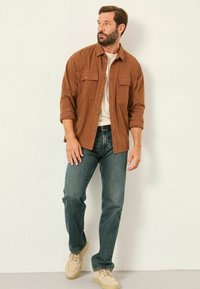 Braun kariertes Hemd, helles T-Shirt darunter, blaue Jeans und beigefarbene Sneakers. Modell steht vor einem schlichten Hintergrund.