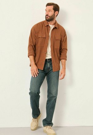 Braun kariertes Hemd, helles T-Shirt darunter, blaue Jeans und beigefarbene Sneakers. Modell steht vor einem schlichten Hintergrund.