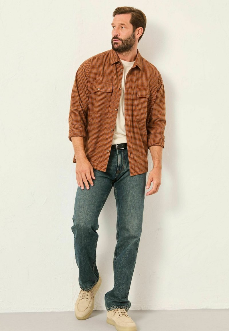 Braun kariertes Hemd, helles T-Shirt darunter, blaue Jeans und beigefarbene Sneakers. Modell steht vor einem schlichten Hintergrund.