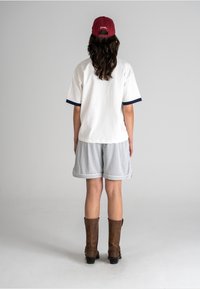 White short-sleeve shirt with navy accents, grey mesh shorts, and brown knee-high boots, with long dark hair under a red cap.