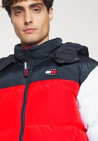 Chaqueta acolchada en negro, rojo y blanco. Presenta un collar alto, cierre de cremallera y parche de logo en el pecho. Material texturizado con acabado brillante.