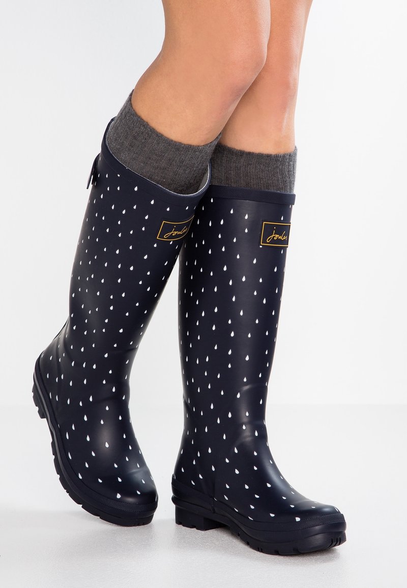 Bottes de pluie bleu marine avec un motif de gouttes de pluie blanches, portées avec des chaussettes grises, montrées sur des jambes croisées contre un fond blanc.