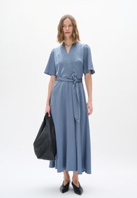 Blaue Wickelkleid mit kurzen Ärmeln, strukturiertem Stoff, seitlichem Bindeband und fließendem Saum, kombiniert mit einer schwarzen Lederhandtasche und spitzen Schuhen.