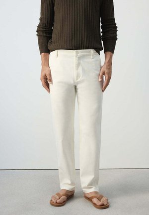Persona que lleva pantalones blancos de pierna recta, un suéter de punto acanalado marrón oscuro y sandalias abiertas marrones, de pie sobre un suelo gris.