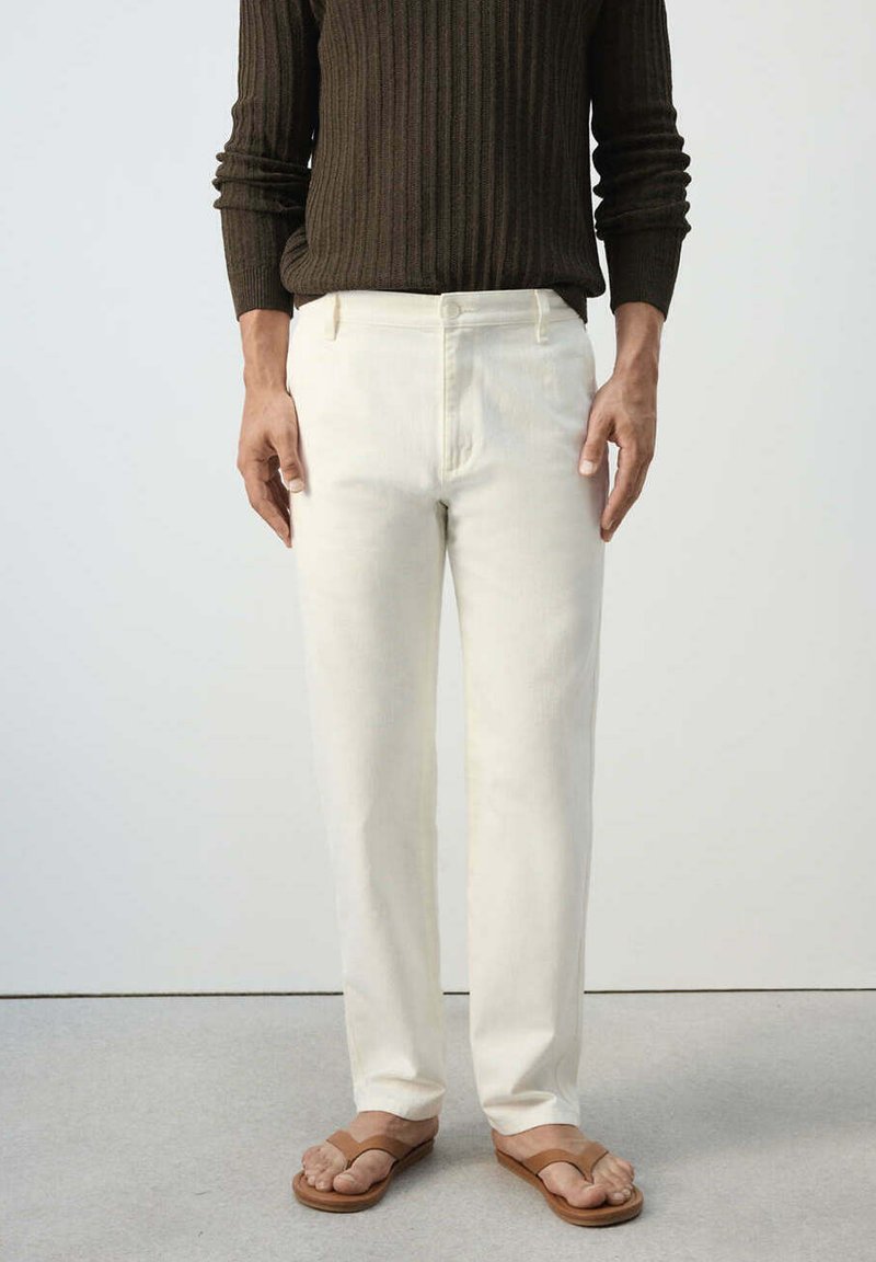 Personne portant un pantalon blanc droit, un pull côtelé marron foncé et des sandales ouvertes beiges, debout sur un sol gris.