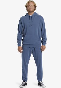 Felpa blu con tasca frontale e coulisse, abbinata a pantaloni della tuta tapered coordinati e scarpe nere. Tessuto in morbido cotone misto.