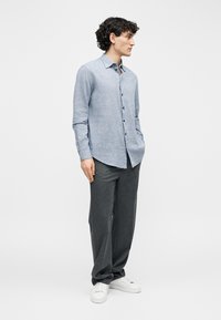 Άνδρας που στέκεται με τα χέρια στις τσέπες, φορώντας ένα ανοιχτό μπλε πουκάμισο με κουμπιά, σκούρο γκρι παντελόνι και λευκά αθλητικά παπούτσια σε απλό φόντο.