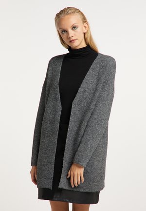 Jonge vrouw draagt een losse grijze cardigan over een zwarte coltrui en zwarte rok, staand tegen een effen lichte achtergrond.
